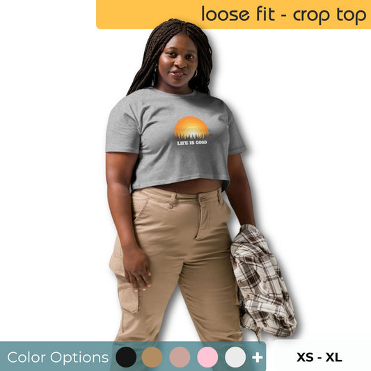 Woman wearing a grey crop top with 'LIFE IS GOOD' and sunset graphic, paired with khaki cargo pants, showcasing color options and available in sizes XS to XL.