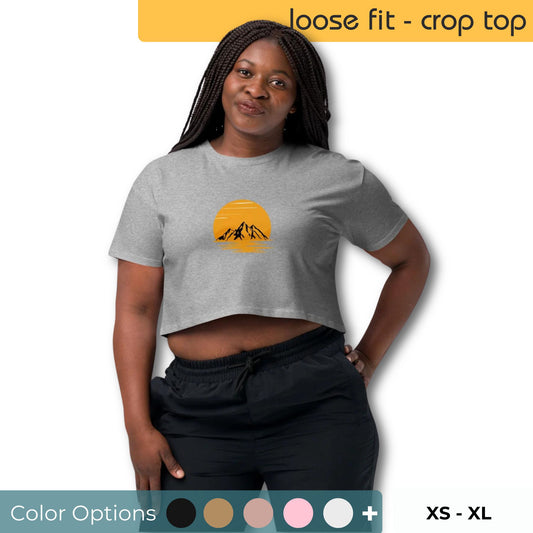 Woman wearing a grey loose-fit crop top with a sunset and mountain graphic design, indicating available color and size options from XS to XL