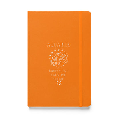 A orange hardcover notebook with 'AQUARIUS' and symbolic illustrations printed in white, along with the words 'INDEPENDENT CREATIVE SOCIAL'. The notebook features an elastic closure.