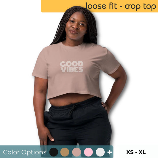 Confident woman modeling a hazy pink loose-fit crop top with 'GOOD VIBES' print, shown with a variety of color swatches and available sizes XS through XL.