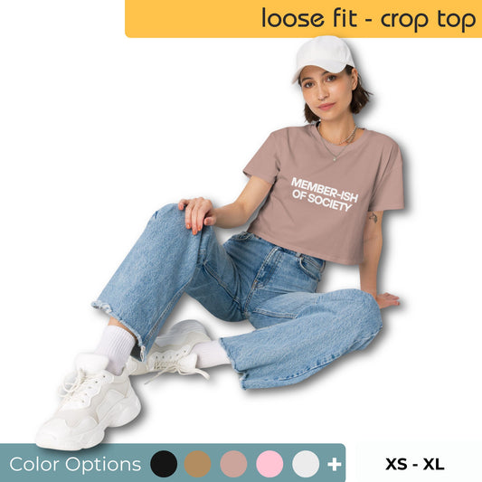 Stylish woman lounging in a taupe crop top with 'MEMBER-ISH OF SOCIETY' text, paired with loose blue jeans and white sneakers, complete with a white cap and displayed color options for a trendy, relaxed look.