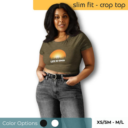 Woman in an olive green slim fit crop top with "Life Is Good" sunrise graphic, paired with casual high-waisted jeans, available in sizes XS/SM to M/L.