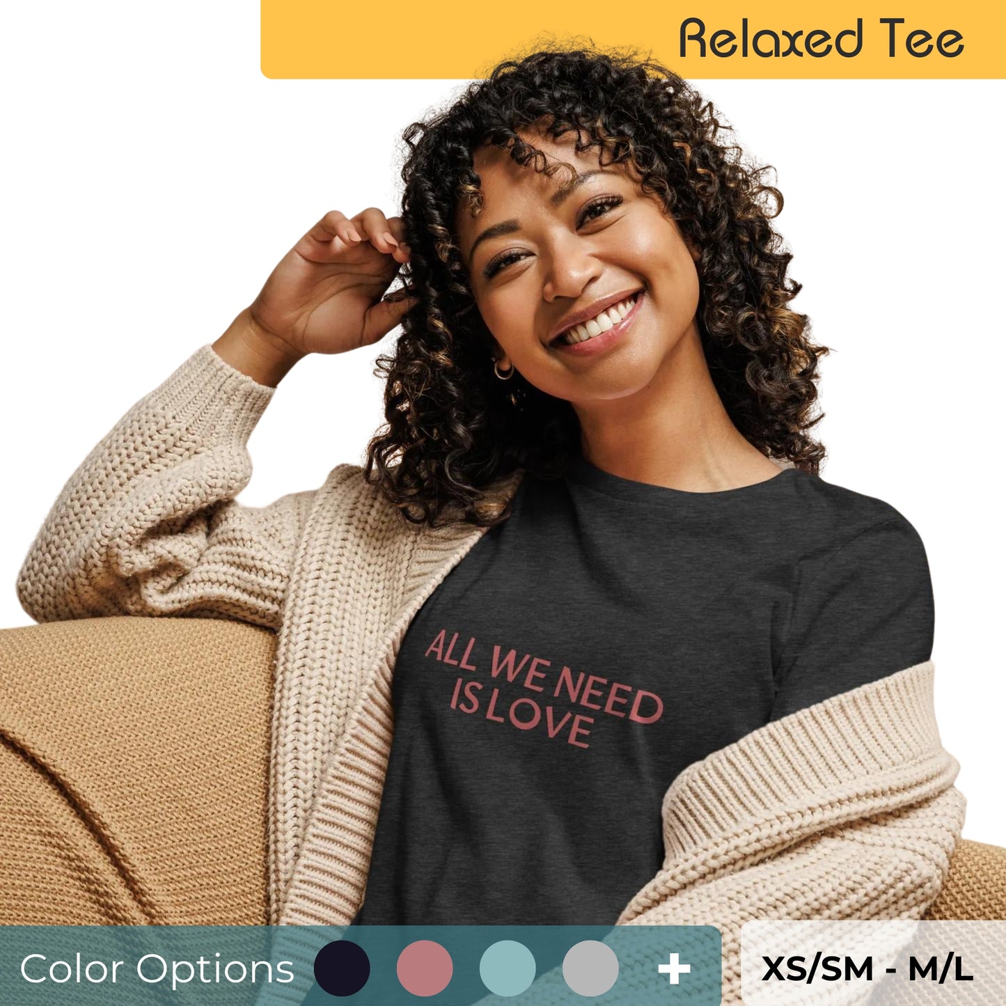 Joyful woman with curly hair in a relaxed fit dark grey tee featuring the phrase 'All We Need Is Love' in red lettering, paired with a beige knitted cardigan, available in multiple colors, sizes XS/SM to M/L.