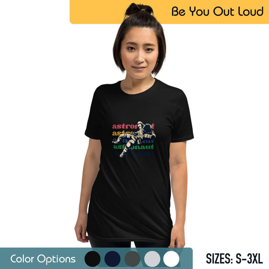 A woman with her hair up in a bun wearing a black T-shirt that has a colorful graphic of an astronaut and the word "astronaut" repeated in a circular pattern. The background features the text "Be You Out Loud," along with a series of color options and the available sizes from S to 3XL.