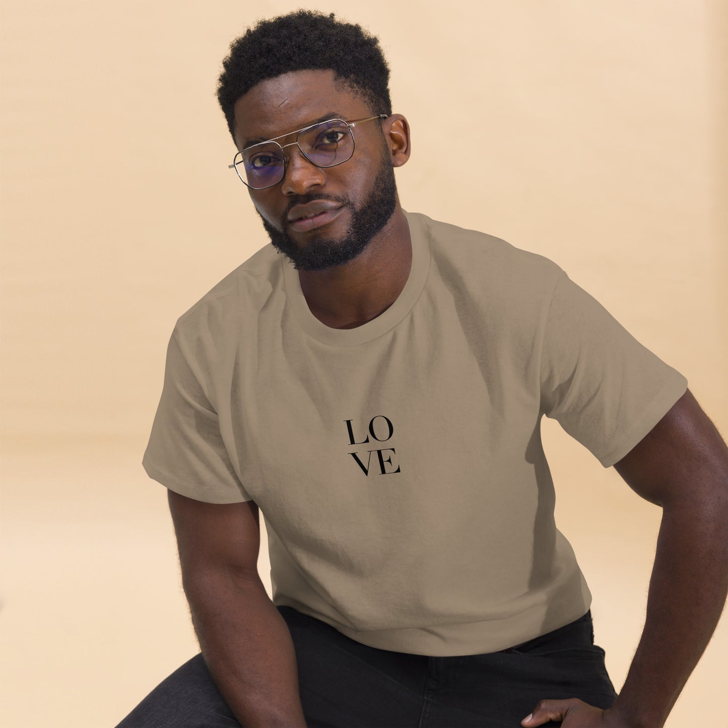 Man in savana sand t-shirt with "LOVE" print leaning forward against a light background, portraying casual style and positive messaging.