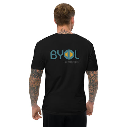 A young man with short blonde hair and arm tattoos wearing a black "BYOL: a mindset" T-shirt, viewed from the back. The T-shirt features the "BYOL" logo in teal and yellow on the back and is available in multiple colors (red, blue, gray, white, plus more) and sizes (S-3XL). The "be you out loud" logo is displayed at the top.