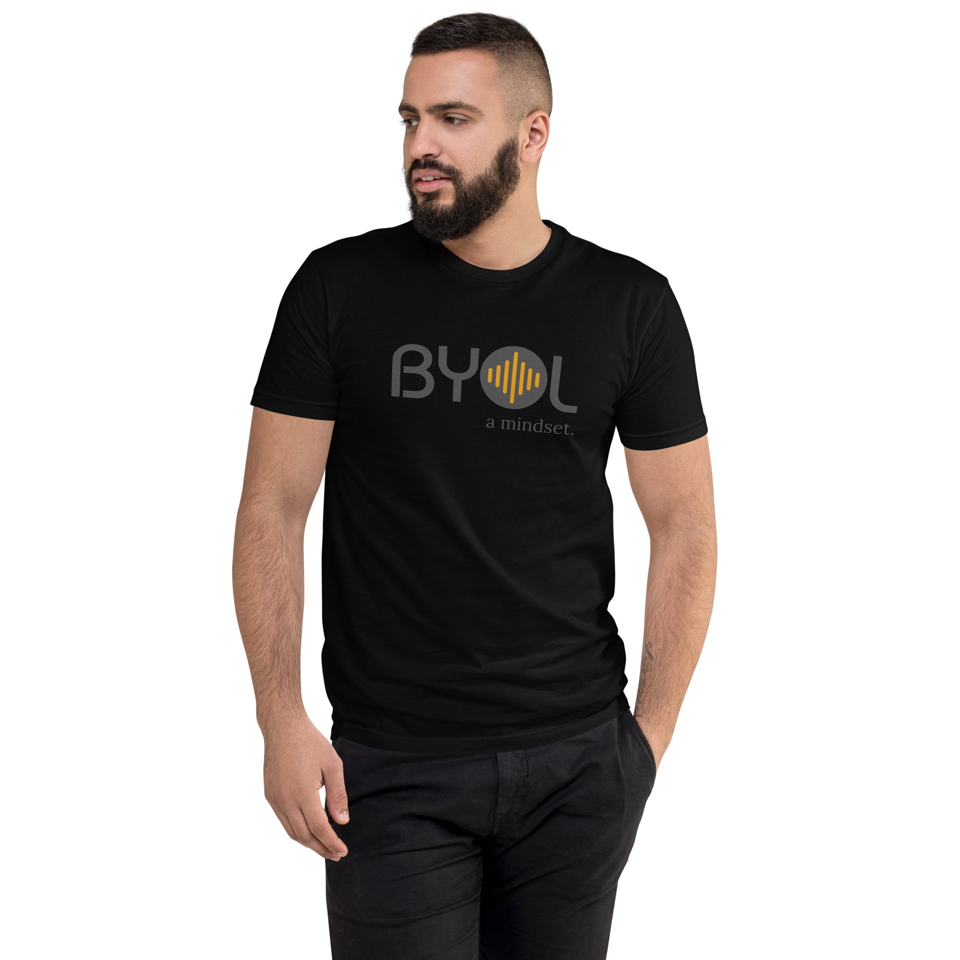 A young man with short dark hair and a beard wearing a black "BYOL: a mindset" T-shirt, standing with one hand in his pocket and looking to the side. The T-shirt features the "BYOL" logo in gray and yellow on the front and is available in multiple colors (red, blue, gray, plus more) and sizes (S-3XL). The "be you out loud" logo is displayed at the top.