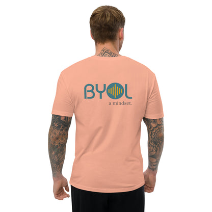 A young man with short blonde hair and arm tattoos wearing a dessert pink "BYOL: a mindset" T-shirt, viewed from the back. The T-shirt features the "BYOL" logo in teal and yellow on the back and is available in multiple colors (red, blue, gray, white, plus more) and sizes (S-3XL). The "be you out loud" logo is displayed at the top.