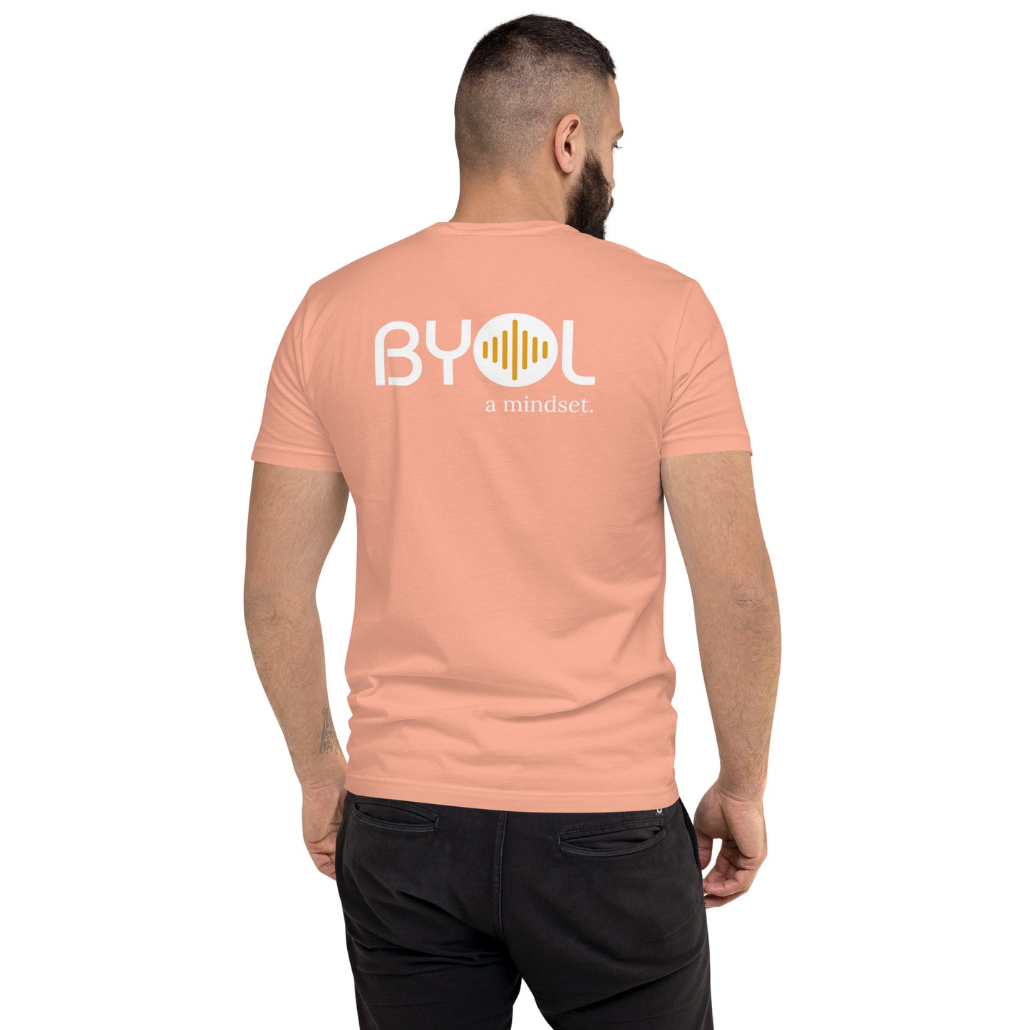 A young man with short dark hair and a beard wearing a dessert pink "BYOL: a mindset" T-shirt, viewed from the back. The T-shirt features the "BYOL" logo in white and yellow on the back and is available in multiple colors (red, blue, gray, white, plus more) and sizes (S-3XL). The "be you out loud" logo is displayed at the top.