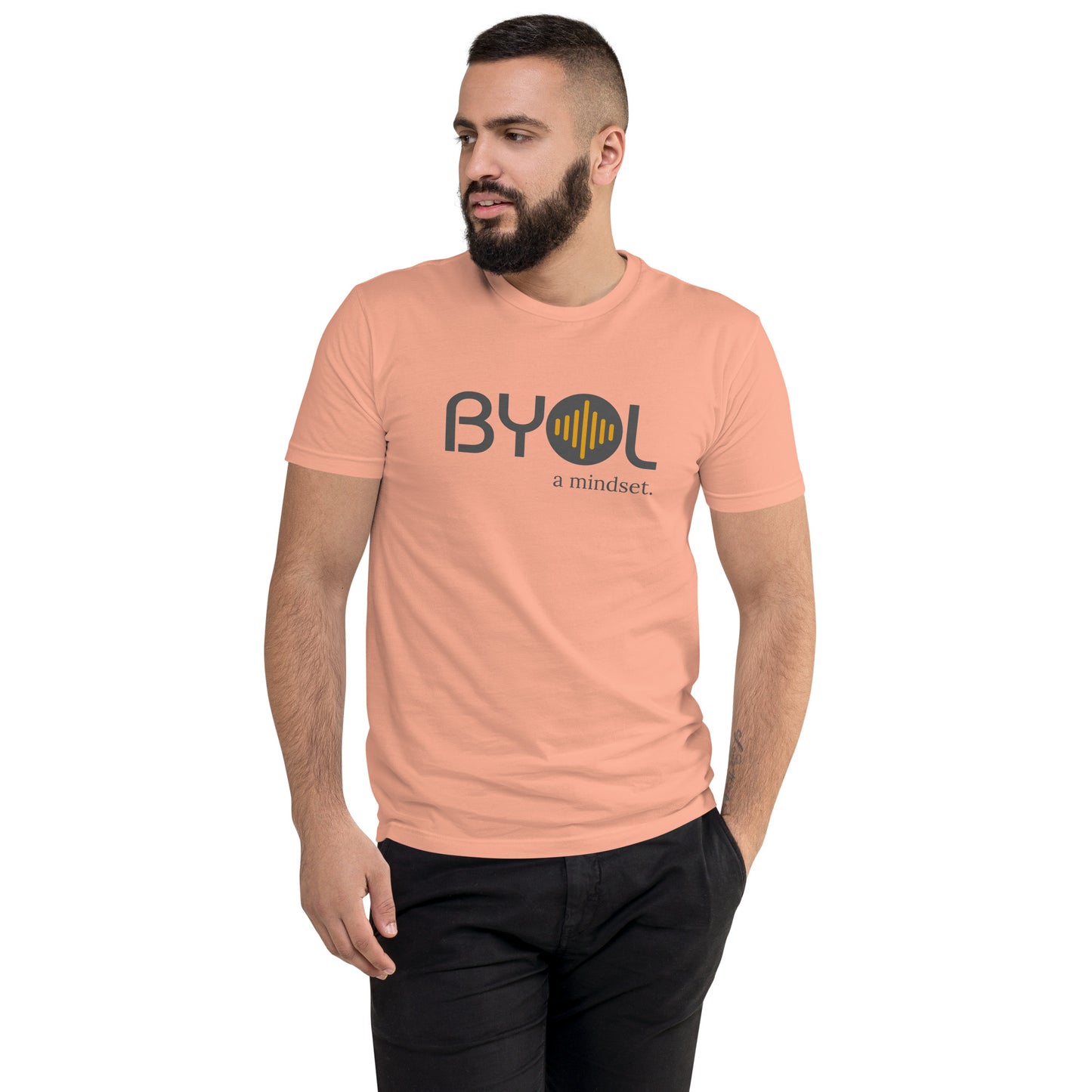 A young man with short dark hair and a beard wearing a dessert pink "BYOL: a mindset" T-shirt, standing with one hand in his pocket and looking to the side. The T-shirt features the "BYOL" logo in gray and yellow on the front and is available in multiple colors (red, blue, gray, plus more) and sizes (S-3XL). The "be you out loud" logo is displayed at the top.