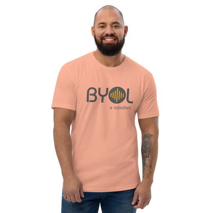 A bearded man with a shaved head wearing a dessert pink "BYOL: a mindset" T-shirt, smiling and standing with his arms at his sides. The T-shirt features the "BYOL" logo in gray and yellow on the front and is available in multiple colors (red, blue, gray, white, plus more) and sizes (S-3XL). The "be you out loud" logo is displayed at the top.