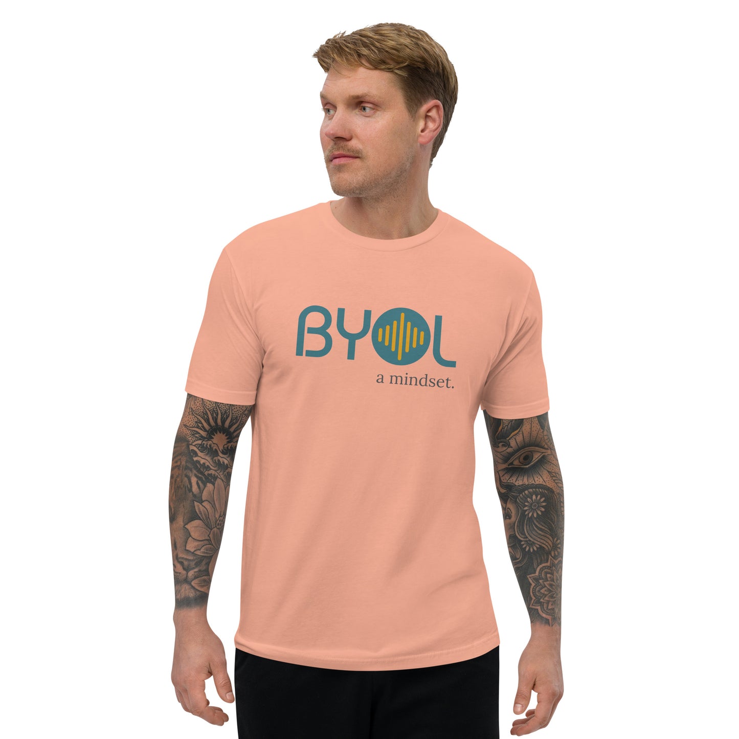 A young man with short blonde hair and arm tattoos wearing a dessert pink "BYOL: a mindset" T-shirt, standing and looking to the side. The T-shirt features the "BYOL" logo in teal and yellow on the front and is available in multiple colors (red, blue, gray, white, plus more) and sizes (S-3XL). The "be you out loud" logo is displayed at the top.