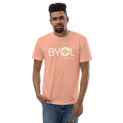 A young man with short curly hair wearing a dessert pink "BYOL: a mindset" T-shirt, standing with one hand in his pocket. The T-shirt features the "BYOL" logo in white and yellow on the front and is available in multiple colors (red, blue, gray, white, plus more) and sizes (S-3XL). The "be you out loud" logo is displayed at the top.