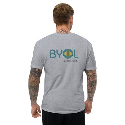 A young man with short blonde hair and arm tattoos wearing a heather grey "BYOL: a mindset" T-shirt, viewed from the back. The T-shirt features the "BYOL" logo in teal and yellow on the back and is available in multiple colors (red, blue, gray, white, plus more) and sizes (S-3XL). The "be you out loud" logo is displayed at the top.