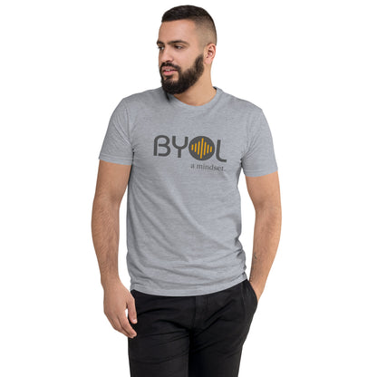 A young man with short dark hair and a beard wearing a heather grey "BYOL: a mindset" T-shirt, standing with one hand in his pocket and looking to the side. The T-shirt features the "BYOL" logo in gray and yellow on the front and is available in multiple colors (red, blue, gray, plus more) and sizes (S-3XL). The "be you out loud" logo is displayed at the top.