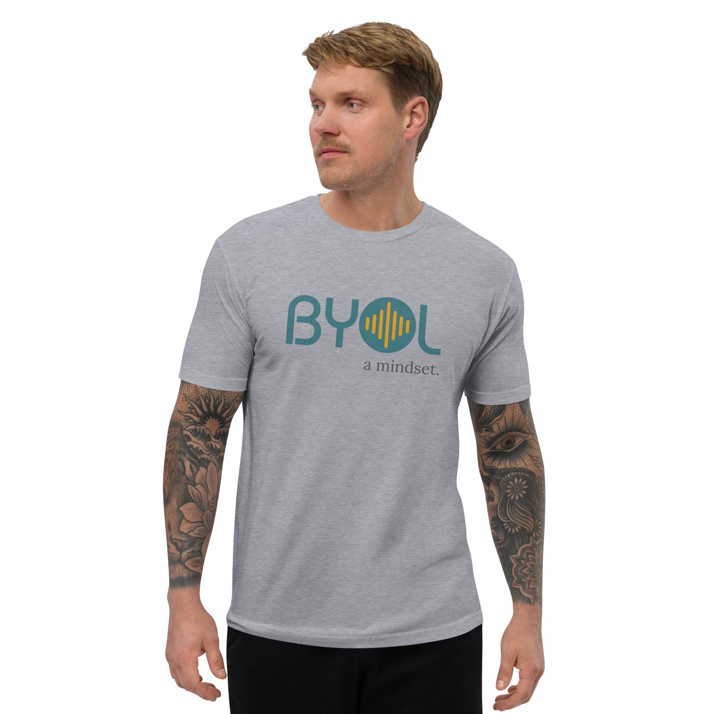 A young man with short blonde hair and arm tattoos wearing a heather grey "BYOL: a mindset" T-shirt, standing and looking to the side. The T-shirt features the "BYOL" logo in teal and yellow on the front and is available in multiple colors (red, blue, gray, white, plus more) and sizes (S-3XL). The "be you out loud" logo is displayed at the top.