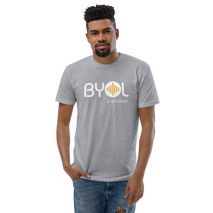 A young man with short curly hair wearing a heather grey "BYOL: a mindset" T-shirt, standing with one hand in his pocket. The T-shirt features the "BYOL" logo in white and yellow on the front and is available in multiple colors (red, blue, gray, white, plus more) and sizes (S-3XL). The "be you out loud" logo is displayed at the top.