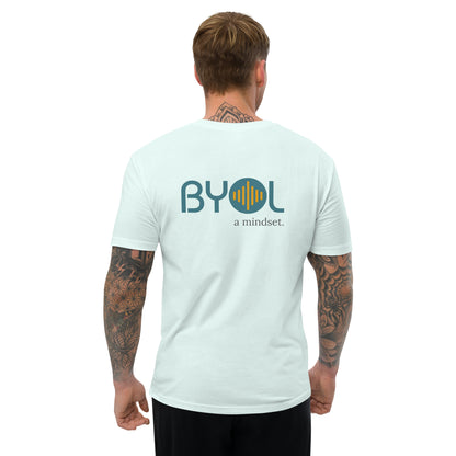 A young man with short blonde hair and arm tattoos wearing a light blue "BYOL: a mindset" T-shirt, viewed from the back. The T-shirt features the "BYOL" logo in teal and yellow on the back and is available in multiple colors (red, blue, gray, white, plus more) and sizes (S-3XL). The "be you out loud" logo is displayed at the top.