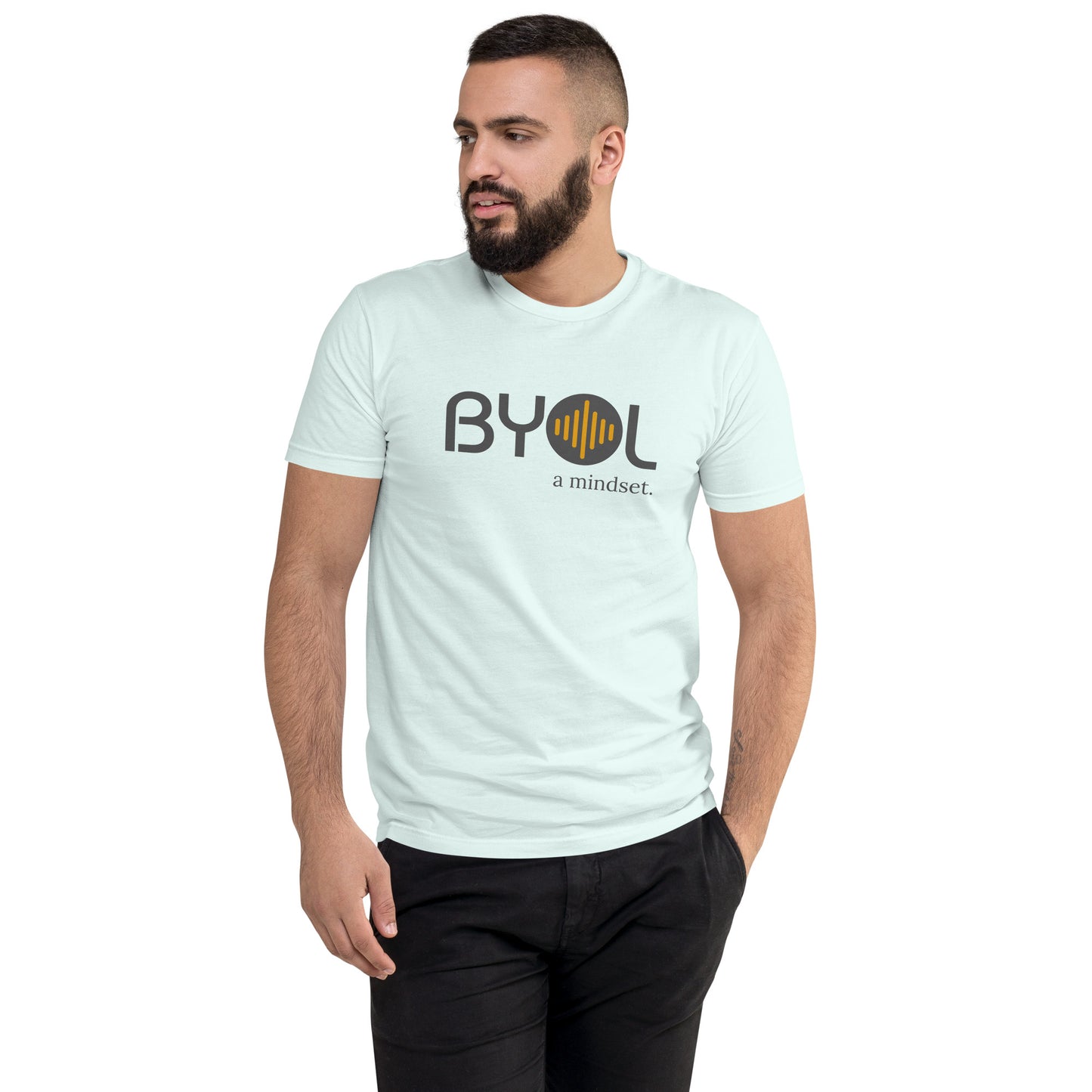 A young man with short dark hair and a beard wearing a light blue "BYOL: a mindset" T-shirt, standing with one hand in his pocket and looking to the side. The T-shirt features the "BYOL" logo in gray and yellow on the front and is available in multiple colors (red, blue, gray, plus more) and sizes (S-3XL). The "be you out loud" logo is displayed at the top.