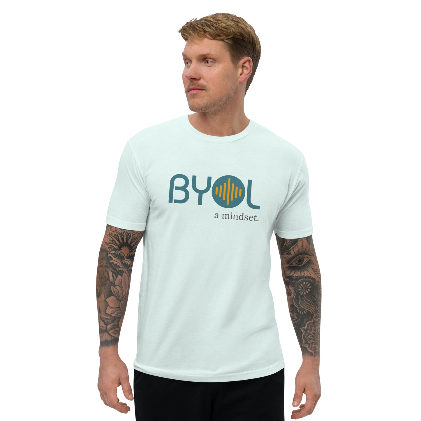 A young man with short blonde hair and arm tattoos wearing a light blue "BYOL: a mindset" T-shirt, standing and looking to the side. The T-shirt features the "BYOL" logo in teal and yellow on the front and is available in multiple colors (red, blue, gray, white, plus more) and sizes (S-3XL). The "be you out loud" logo is displayed at the top.