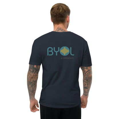 A young man with short blonde hair and arm tattoos wearing a navy blue "BYOL: a mindset" T-shirt, viewed from the back. The T-shirt features the "BYOL" logo in teal and yellow on the back and is available in multiple colors (red, blue, gray, white, plus more) and sizes (S-3XL). The "be you out loud" logo is displayed at the top.