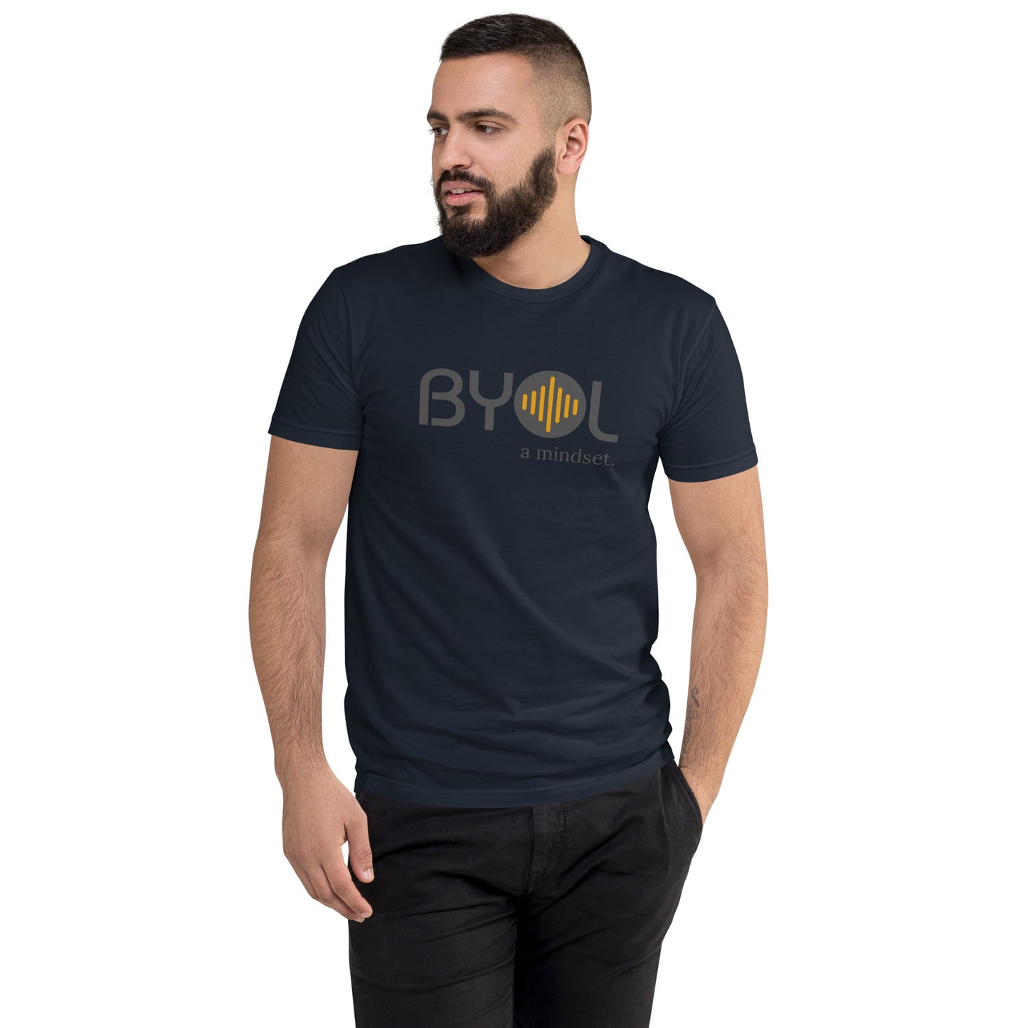 A young man with short dark hair and a beard wearing a navy blue "BYOL: a mindset" T-shirt, standing with one hand in his pocket and looking to the side. The T-shirt features the "BYOL" logo in gray and yellow on the front and is available in multiple colors (red, blue, gray, plus more) and sizes (S-3XL). The "be you out loud" logo is displayed at the top.