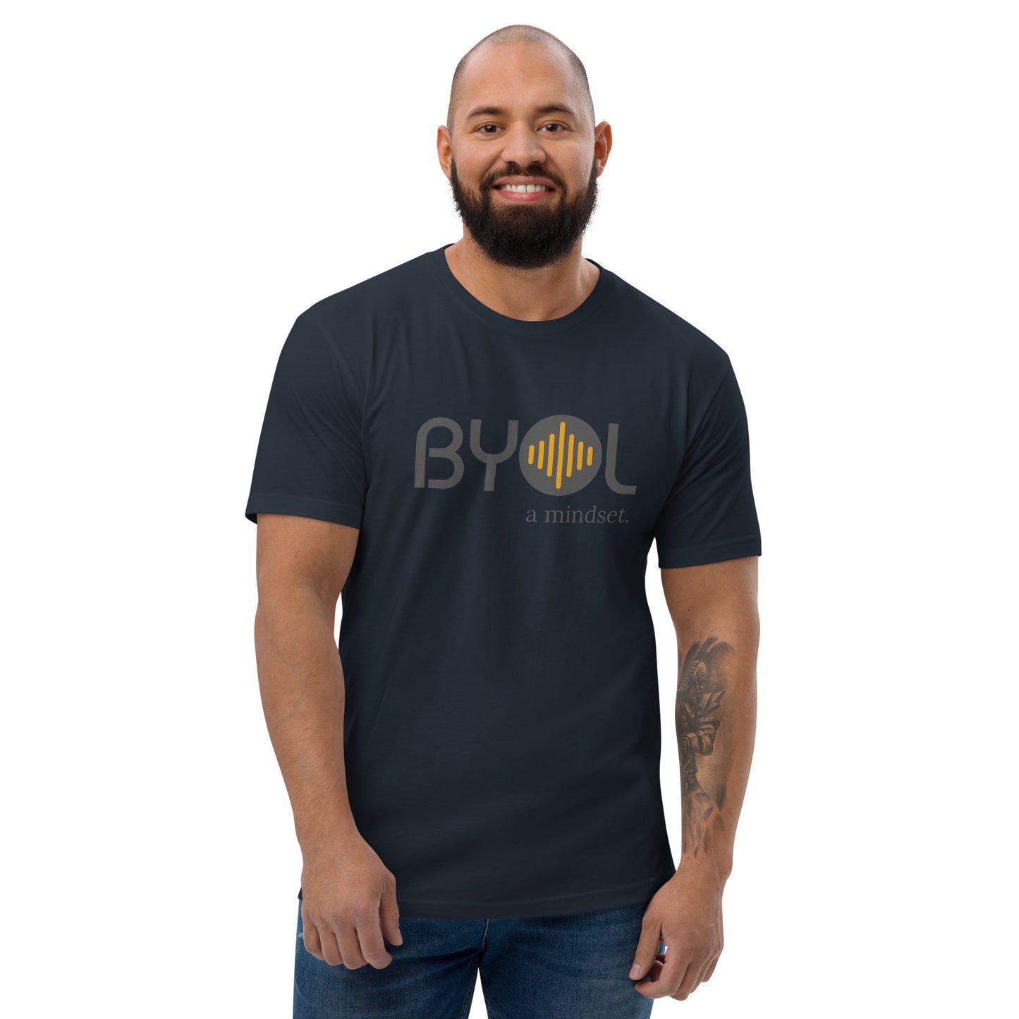 A bearded man with a shaved head wearing a navy blue "BYOL: a mindset" T-shirt, smiling and standing with his arms at his sides. The T-shirt features the "BYOL" logo in gray and yellow on the front and is available in multiple colors (red, blue, gray, white, plus more) and sizes (S-3XL). The "be you out loud" logo is displayed at the top.