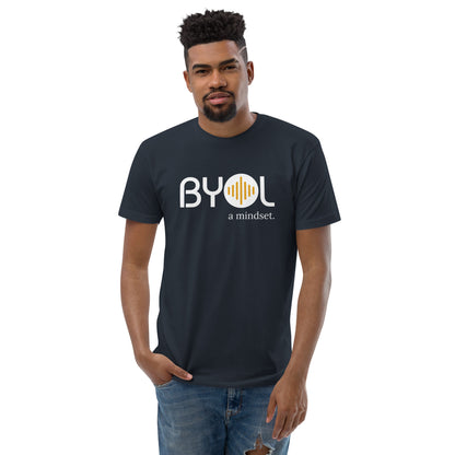 A young man with short curly hair wearing a navy blue "BYOL: a mindset" T-shirt, standing with one hand in his pocket. The T-shirt features the "BYOL" logo in white and yellow on the front and is available in multiple colors (red, blue, gray, white, plus more) and sizes (S-3XL). The "be you out loud" logo is displayed at the top.