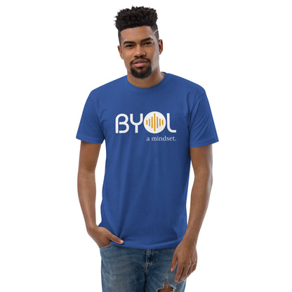 A young man with short curly hair wearing a blue "BYOL: a mindset" T-shirt, standing with one hand in his pocket. The T-shirt features the "BYOL" logo in white and yellow on the front and is available in multiple colors (red, blue, gray, white, plus more) and sizes (S-3XL). The "be you out loud" logo is displayed at the top.