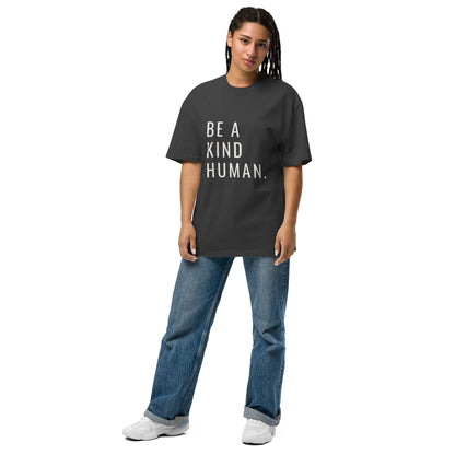 A woman in a casual stance, sporting a faded black oversized t-shirt with "BE A KIND HUMAN." in white block letters, complemented by classic blue jeans and white sneakers.