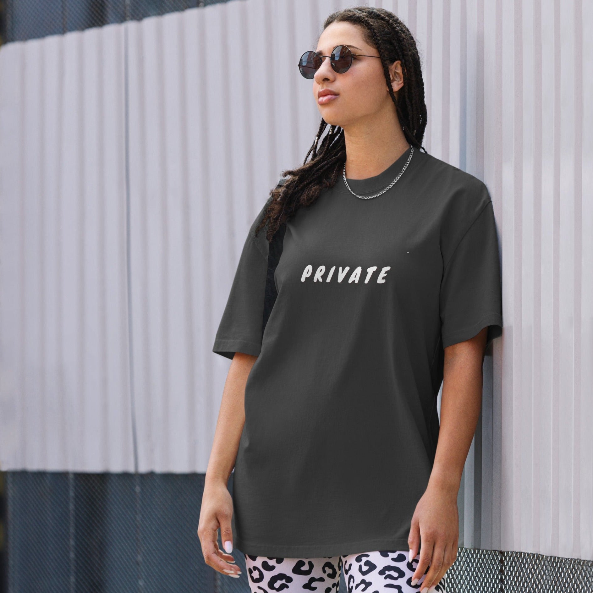 A woman exudes a confident yet understated vibe in an urban setting, wearing a faded black oversized t-shirt with the word "PRIVATE" printed in white across the chest. The simple, bold statement contrasts with her playful white and black leopard print pants. Her style is accessorized with sunglasses and a silver necklace, creating a look that balances a personal statement of discretion with a touch of edgy street fashion.