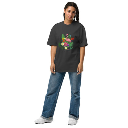 A woman is wearing a faded black oversized t-shirt adorned with a colorful tropical design featuring a pink flamingo surrounded by lush greenery and bright flowers. The playful and vibrant graphic stands out against the tee's dark background. She pairs it with casual blue jeans and white sneakers, presenting a relaxed yet stylish ensemble that evokes a sense of fun and a tropical vibe. Her posture is easygoing, suggesting comfort and a laid-back fashion sense.