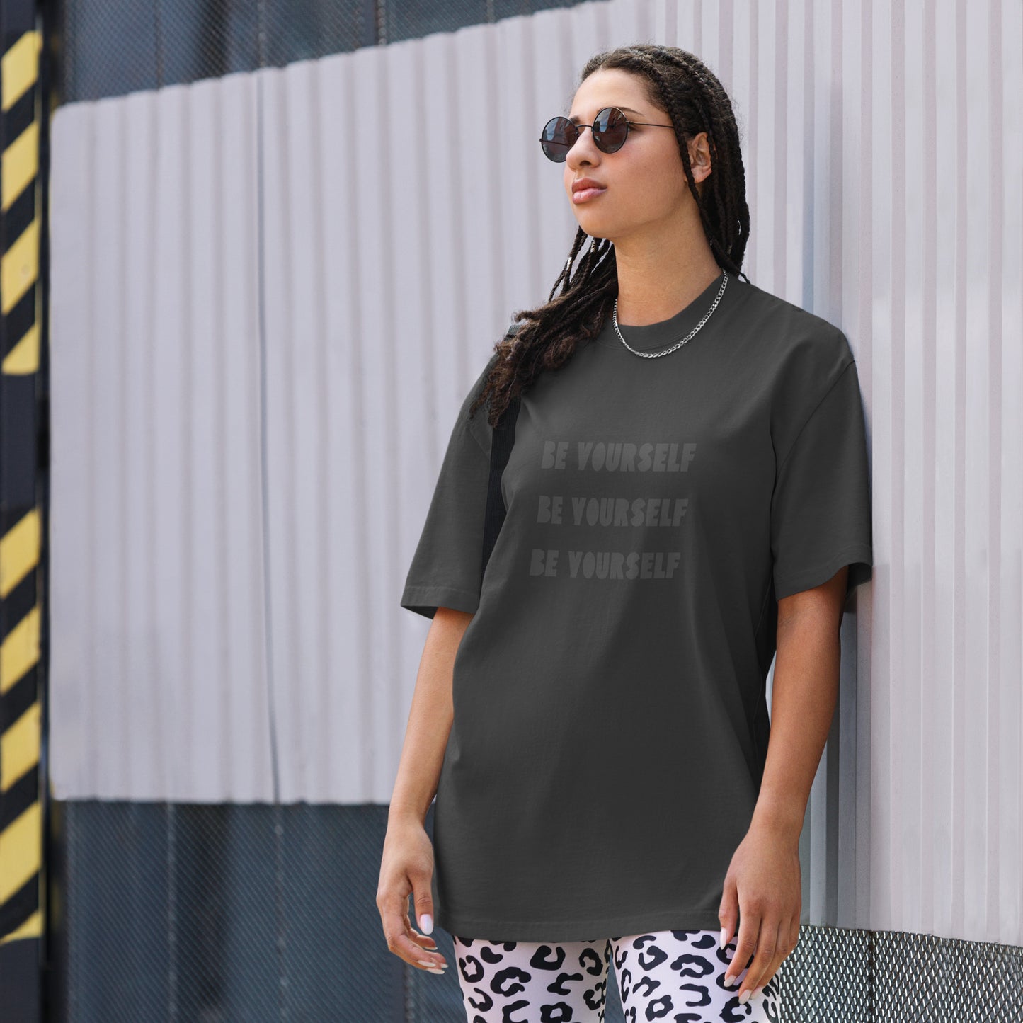 A self-assured woman wears the Women’s ‘Be Yourself’ faded black Oversized Tee leaning g against a wall wearing cheetah stretchy pants and circle sunglasses. 