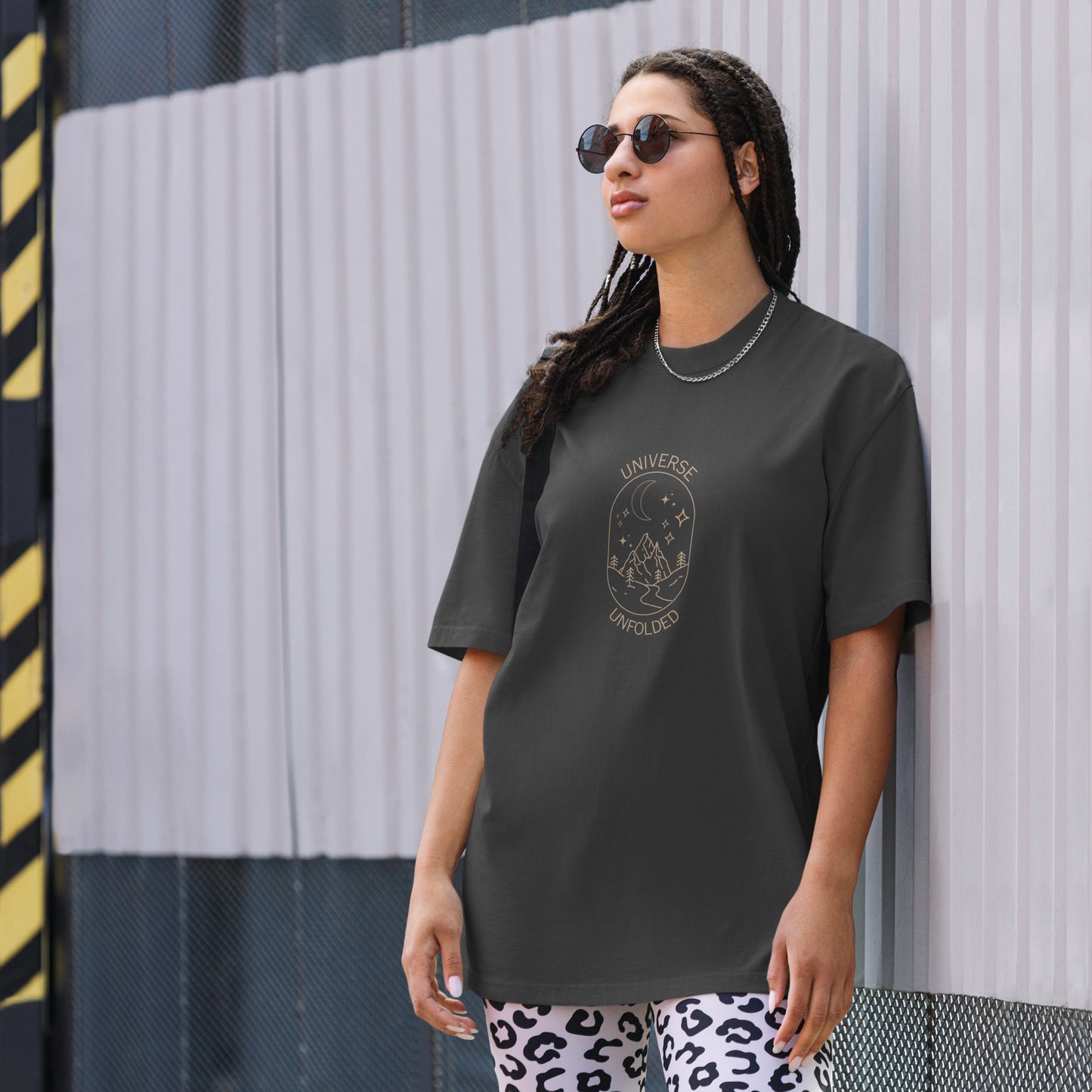 Trendsetting woman wearing a Be You Out Loud Oversized Tee in faded black, featuring a 'Universe Unfolded' graphic design that suggests cosmic exploration and self-discovery, accented with fashion-forward accessories.