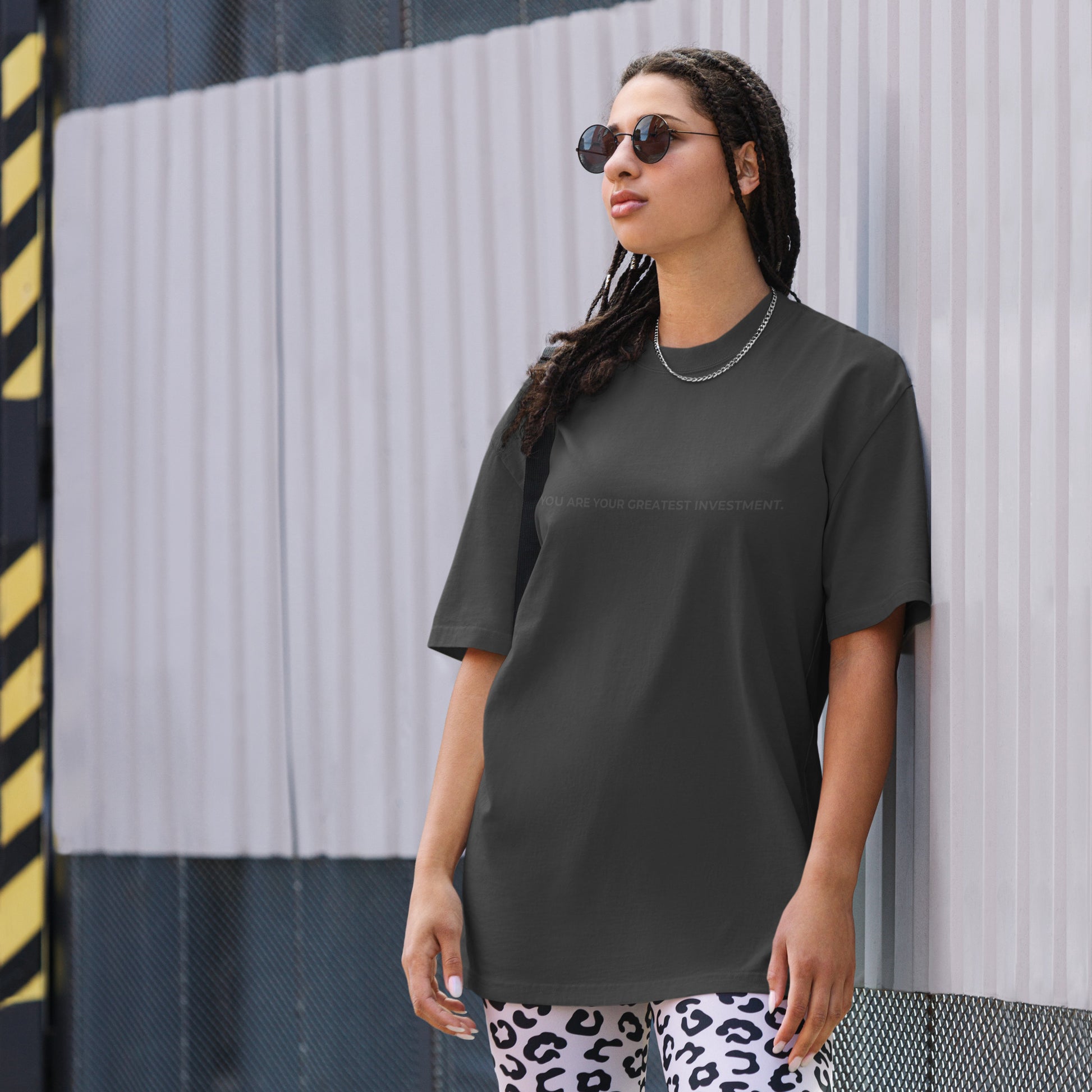 A woman wearing a faded black "You are your greatest investment" t-shirt, styled with leopard print leggings and sunglasses, leaning against a corrugated metal wall with a confident expression. The casual yet trendy outfit showcases self-expression and boldness. Perfect for promoting empowering fashion statements.