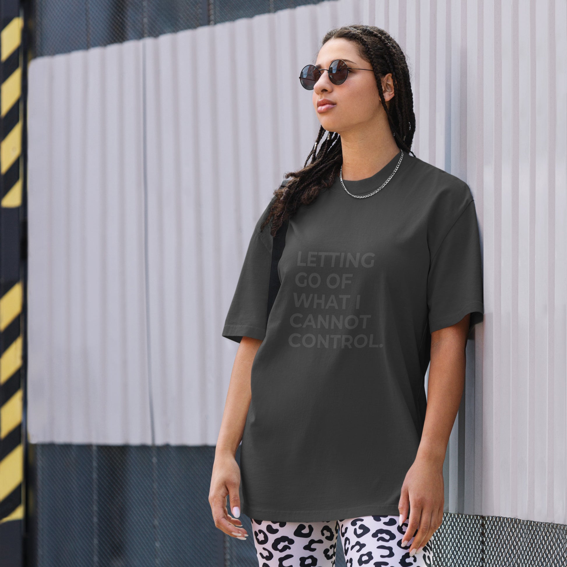 A woman wearing a faded black "Letting go of what I cannot control" t-shirt, styled with leopard print leggings and sunglasses, leaning against a corrugated metal wall with a confident expression. The casual yet trendy outfit showcases self-expression and boldness. Perfect for promoting empowering fashion statements.