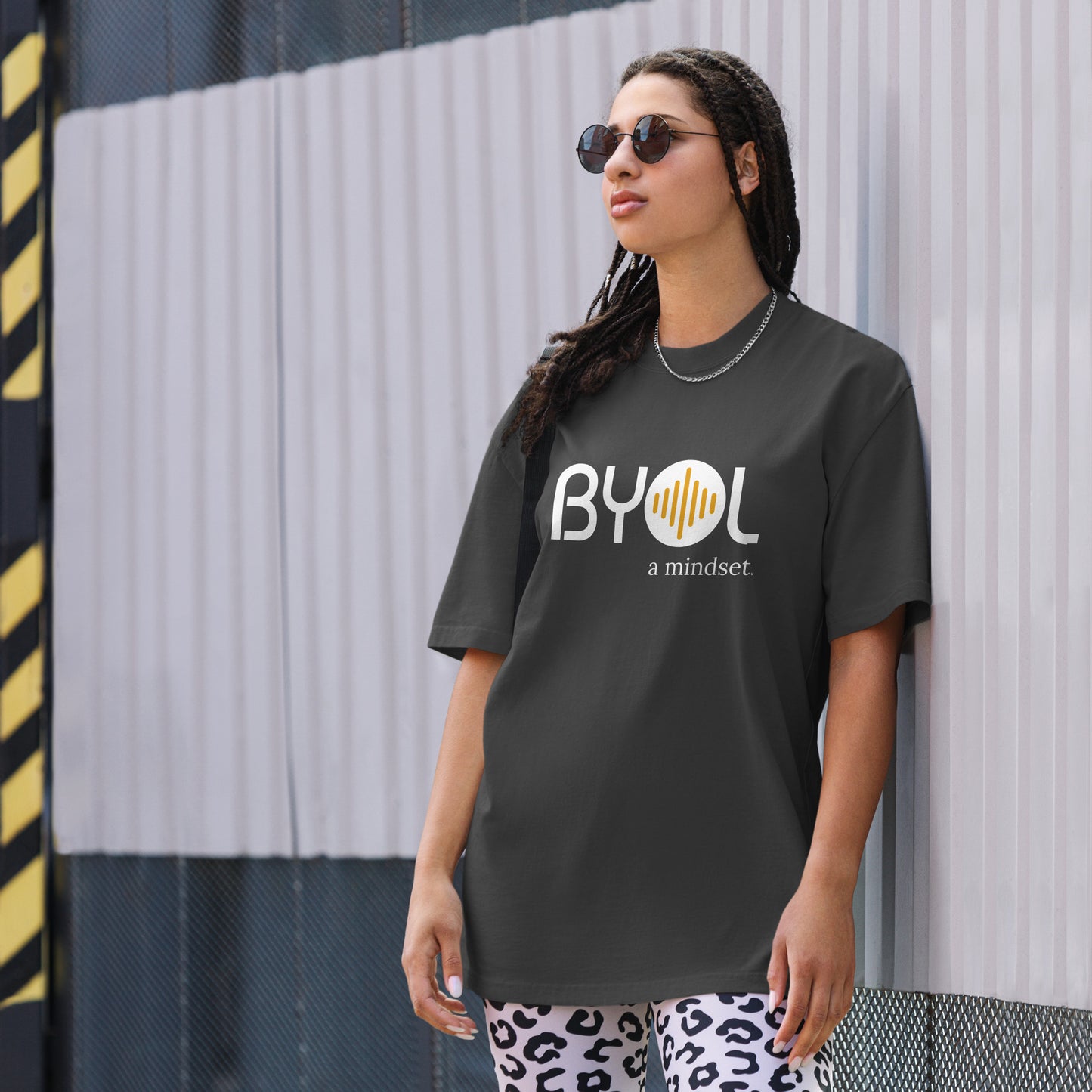 A young woman with long braids wearing sunglasses and a faded black "BYOL: a mindset" T-shirt, standing and looking to the side. The T-shirt features the "BYOL" logo in white and yellow on the front and is available in multiple colors (dark gray, beige, light blue, white, plus more) and sizes (S-3XL). The "be you out loud" logo is displayed at the top.