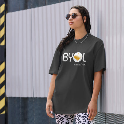 A young woman with long braids wearing sunglasses and a faded black "BYOL: a mindset" T-shirt, standing and looking to the side. The T-shirt features the "BYOL" logo in white and yellow on the front and is available in multiple colors (dark gray, beige, light blue, white, plus more) and sizes (S-3XL). The "be you out loud" logo is displayed at the top.