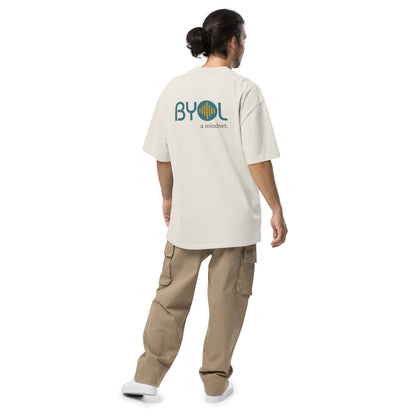 A young man with long hair in a ponytail wearing a faded bone "BYOL: a mindset" T-shirt, viewed from the back. The T-shirt features the "BYOL" logo in teal and yellow on the back and is available in multiple colors (dark gray, beige, light blue) and sizes (S-3XL). The "be you out loud" logo is displayed at the top.