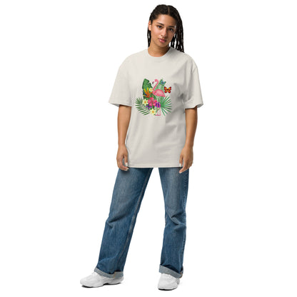 A woman is wearing a stone oversized t-shirt adorned with a colorful tropical design featuring a pink flamingo surrounded by lush greenery and bright flowers. The playful and vibrant graphic stands out against the tee's dark background. She pairs it with casual blue jeans and white sneakers, presenting a relaxed yet stylish ensemble that evokes a sense of fun and a tropical vibe. Her posture is easygoing, suggesting comfort and a laid-back fashion sense.