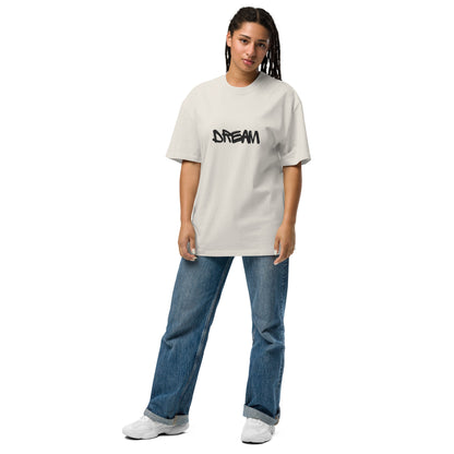 A woman is standing wearing a stone, oversized t-shirt with the word "DREAM" written in black, stylized font across the chest. The tee is complemented with classic blue denim jeans and casual white sneakers, giving off a laid-back yet aspirational vibe. Her relaxed and confident pose suggests a comfortable ease, reflecting a simple, artistic, and dream-inspired style.