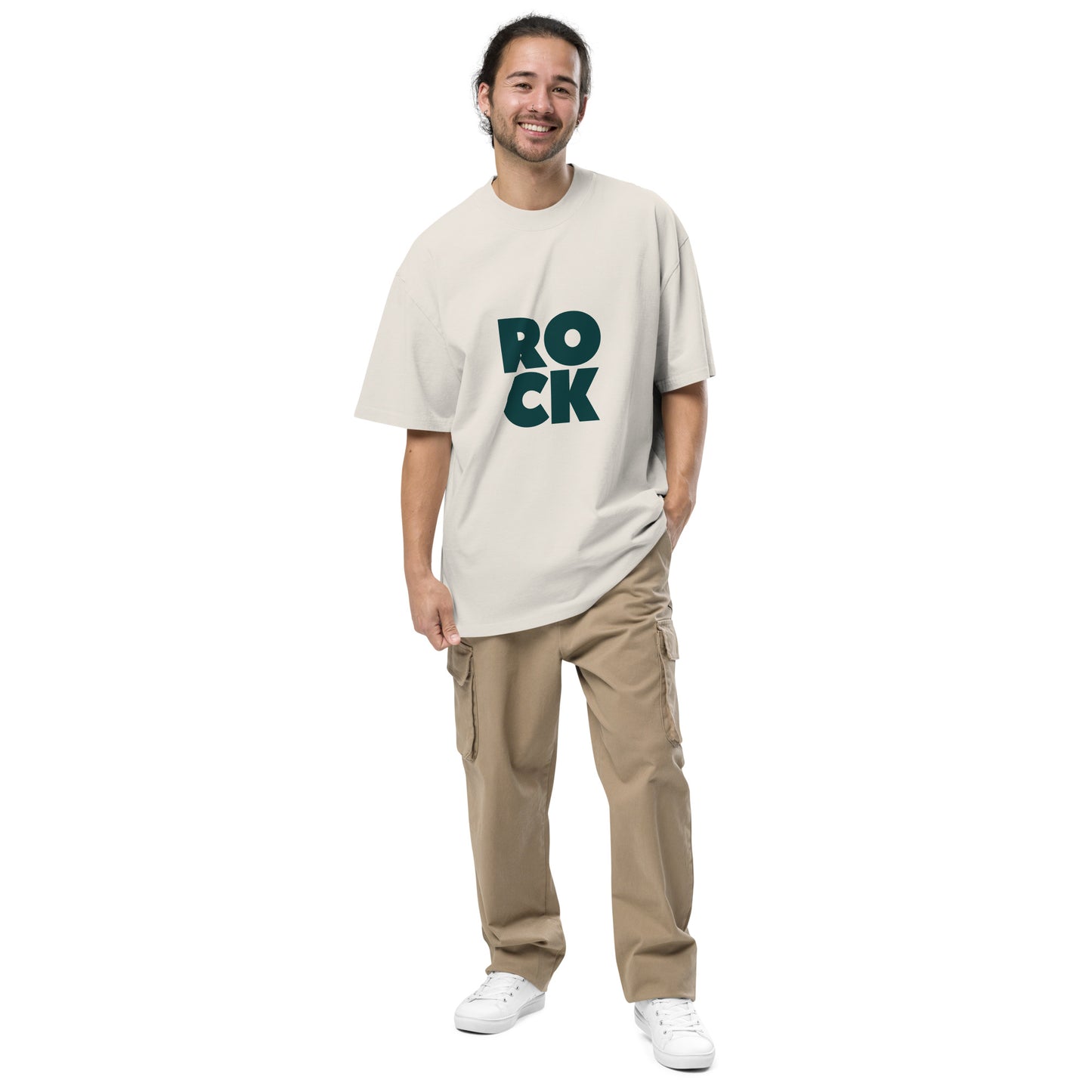 Smiling man wearing a 'Be You Out Loud' tan oversized stone faded t-shirt with bold green 'ROCK' lettering, paired with tan cargo pants and white sneakers