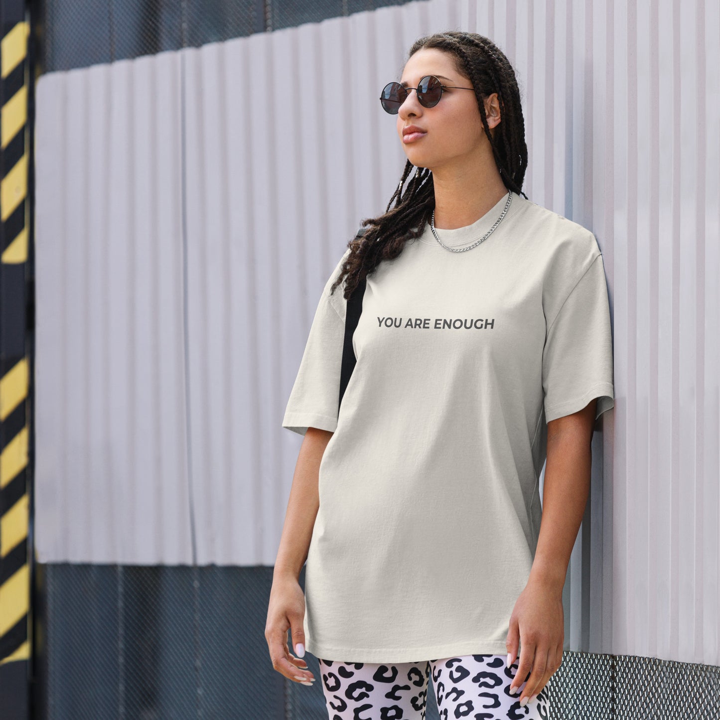 A woman wearing a faded bone "you are enough" t-shirt, styled with leopard print leggings and sunglasses, leaning against a corrugated metal wall with a confident expression. The casual yet trendy outfit showcases self-expression and boldness. Perfect for promoting empowering fashion statements.