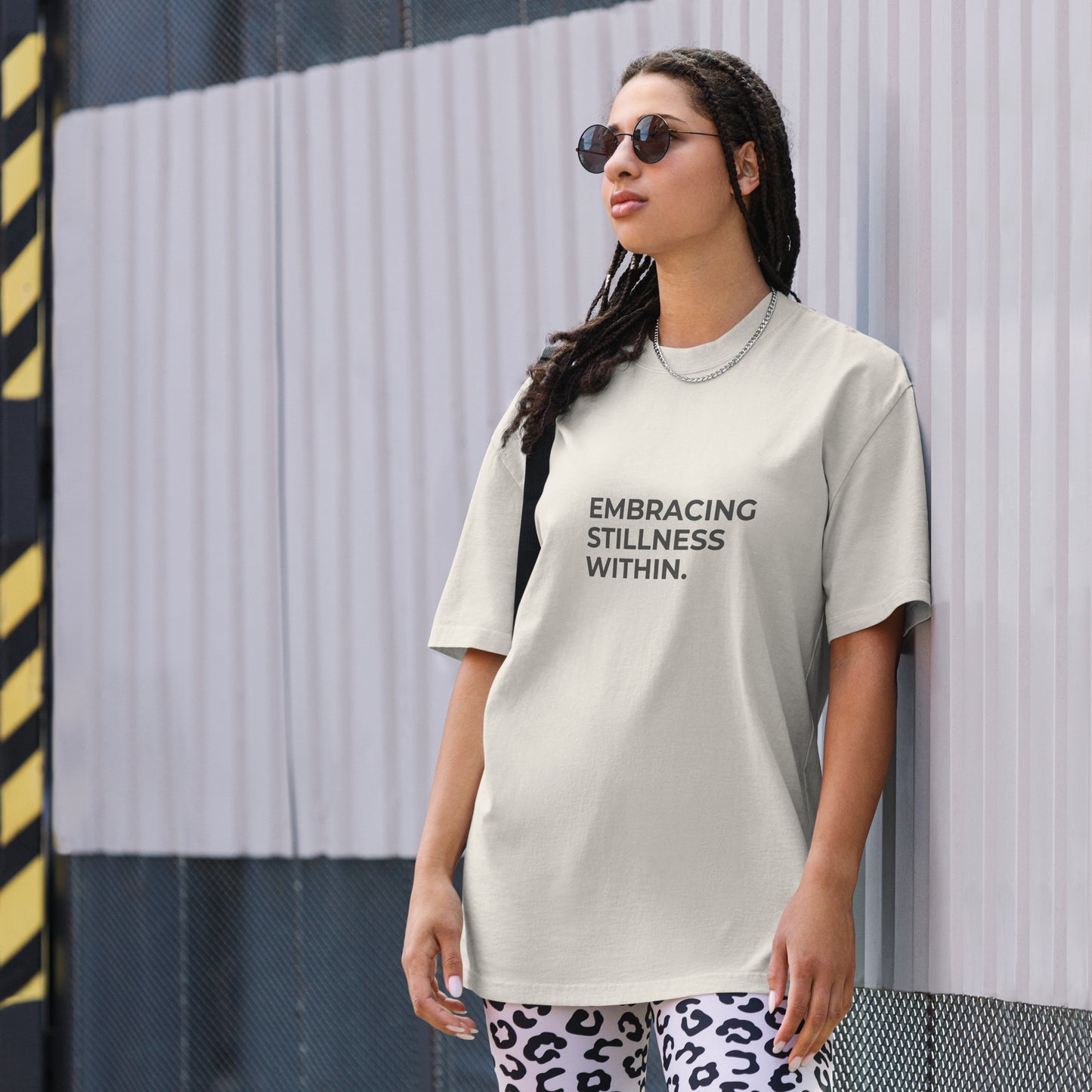 A woman wearing a faded bone "Embracing stillness within" t-shirt, styled with leopard print leggings and sunglasses, leaning against a corrugated metal wall with a confident expression. The casual yet trendy outfit showcases self-expression and boldness. Perfect for promoting empowering fashion statements.