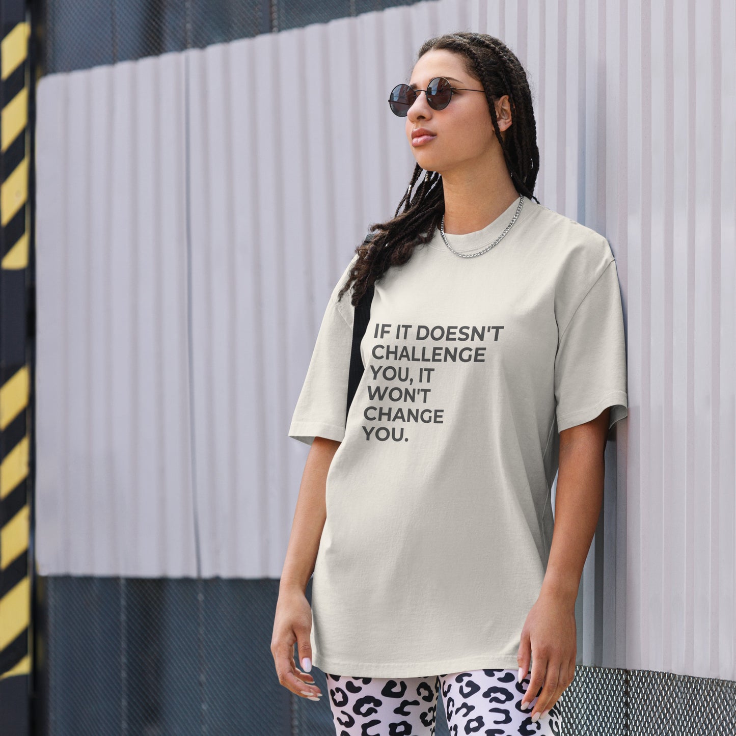 A woman wearing a faded bone "If it doesn't challenge you, it won'f change you" t-shirt, styled with leopard print leggings and sunglasses, leaning against a corrugated metal wall with a confident expression. The casual yet trendy outfit showcases self-expression and boldness. Perfect for promoting empowering fashion statements.