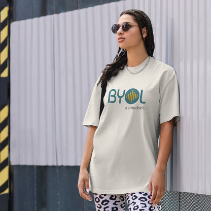 A young woman with long braids wearing sunglasses and a light faded bone "BYOL: a mindset" T-shirt, standing and looking to the side. The T-shirt features the "BYOL" logo in teal and yellow on the front and is available in multiple colors (dark gray, beige, light blue, white, plus more) and sizes (S-3XL). The "be you out loud" logo is displayed at the top.