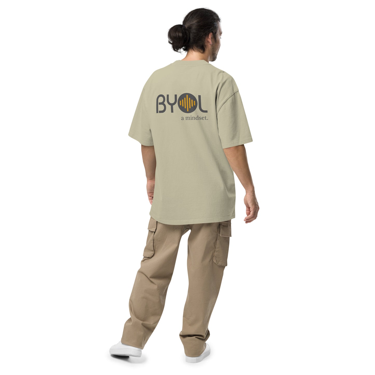 A young man with long hair in a ponytail wearing a eucalyptus "BYOL: a mindset" T-shirt, viewed from the back. The T-shirt features the "BYOL" logo on the back and is available in multiple colors (dark gray, beige, light blue) and sizes (S-3XL). The "be you out loud" logo is displayed at the top.