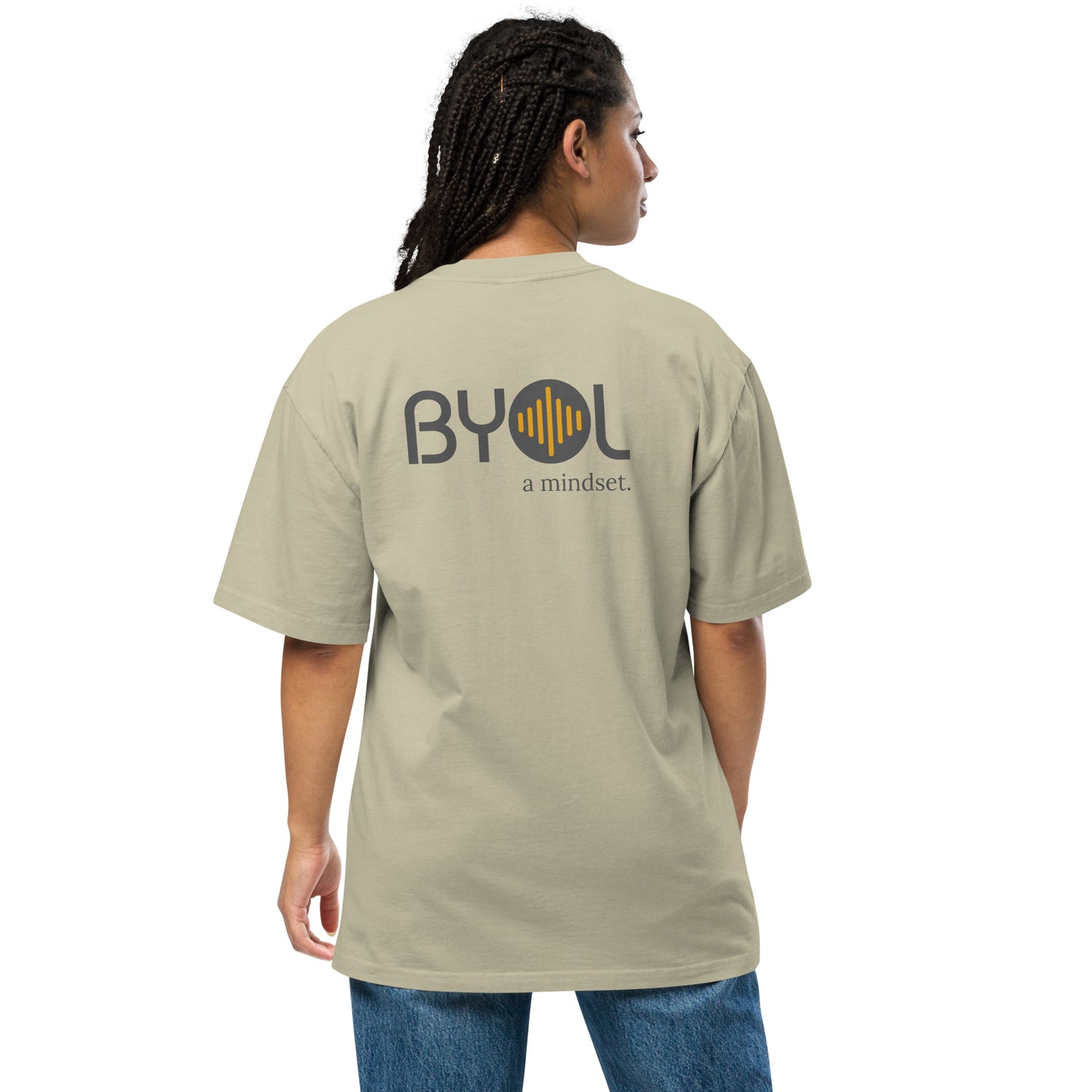 A young woman with long braids wearing a light faded eucalyptus "BYOL: a mindset" T-shirt, viewed from the back. The T-shirt features the "BYOL" logo in dark gray and yellow on the back and is available in multiple colors (dark gray, beige, light blue, white, plus more) and sizes (S-3XL). The "be you out loud" logo is displayed at the top.