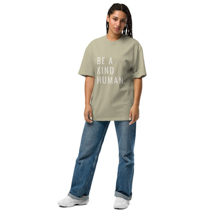 A woman in a casual stance, sporting a faded eucalyptus oversized t-shirt with "BE A KIND HUMAN." in white block letters, complemented by classic blue jeans and white sneakers.