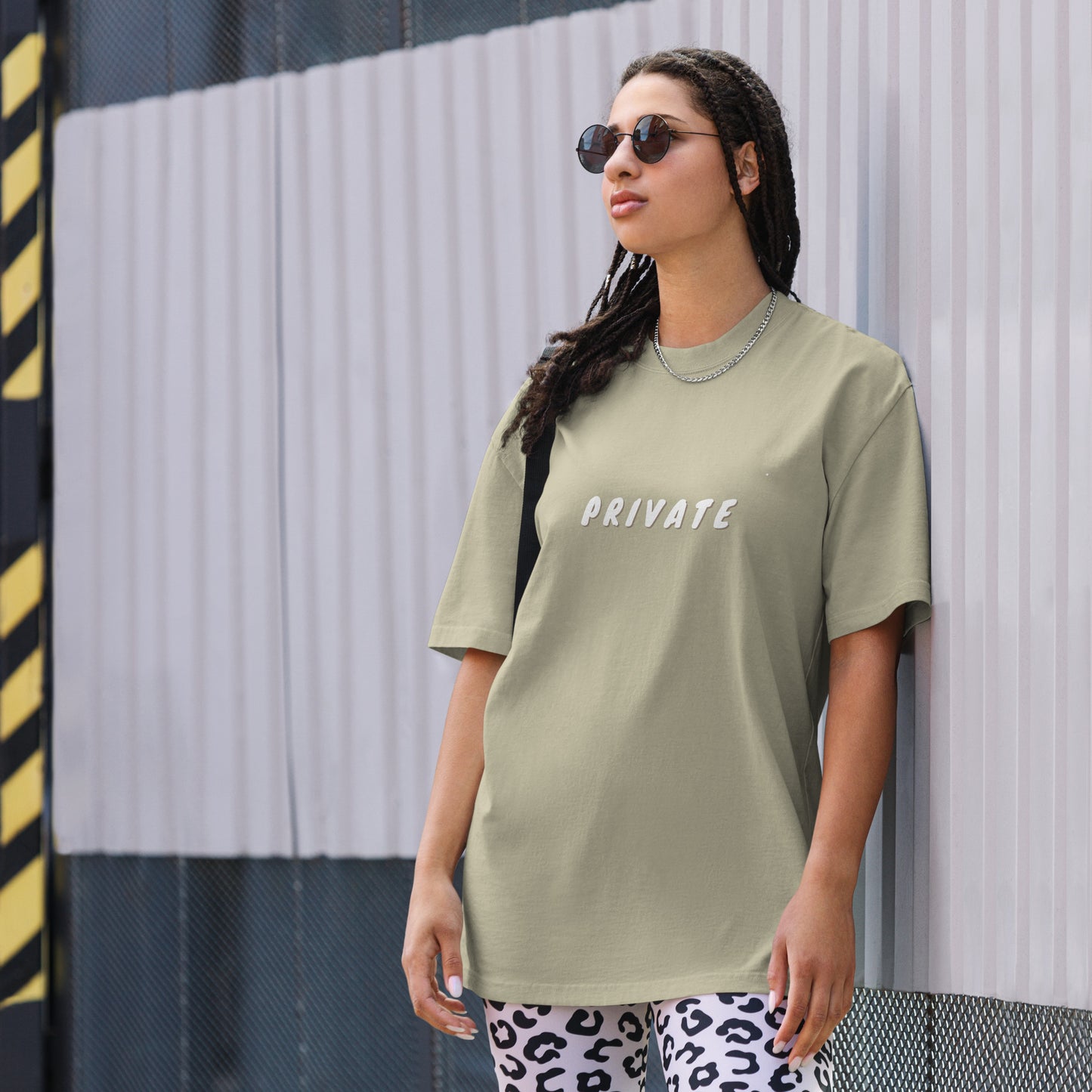 A woman exudes a confident yet understated vibe in an urban setting, wearing a eucalyptus oversized t-shirt with the word "PRIVATE" printed in white across the chest. The simple, bold statement contrasts with her playful white and black leopard print pants. Her style is accessorized with sunglasses and a silver necklace, creating a look that balances a personal statement of discretion with a touch of edgy street fashion.